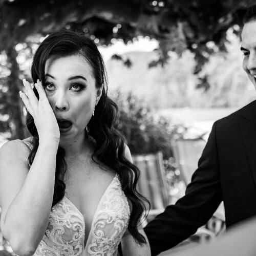 Düğün fotoğrafı: bride surprised expression, groom smiling beside, outdoor garden setting, black and white portrait, dappled tree shade, formal wedding attire | Mischa Baettig, Steinhausen, İsviçre