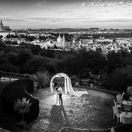 Zdjęcie ślubne: bride and groom embracing, white veil flowing, hilltop garden setting, cathedral in distance, black and white portrait, dramatic cloudy sky | Petr Hrubeš, Brno, Czechy