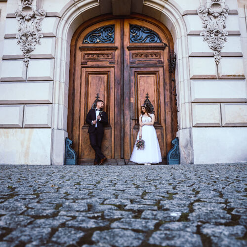 Wedding photo: Silesia | bride and groom at ornate door, couple standing on cobblestone, wooden double doors with carvings, neoclassical architecture with corbels | Marek Kopiec, Katowice, Poland
