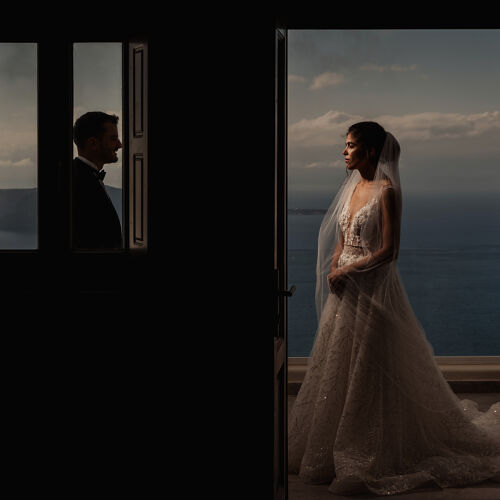 Fotografie de nuntă: bride in lace gown, groom looking through window, couple separated by doorway, golden hour light, sea and sky backdrop | Theodore Vourlis, Athens, Grecia