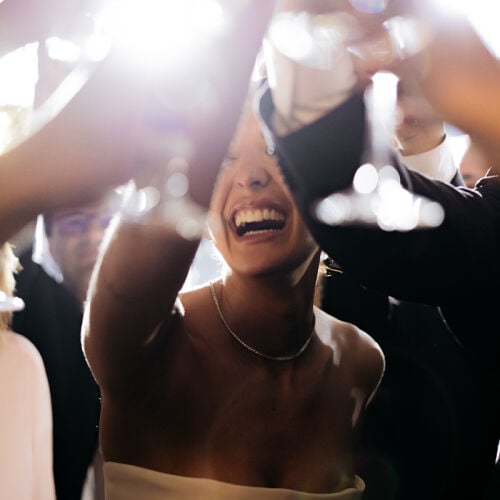 Bryllupsfoto: woman laughing at celebration, bright lights bokeh effect, festive party atmosphere, raised arms dancing, indoor evening event, joyful crowd moment | Eric Corbacho, São Paulo, Brasilien