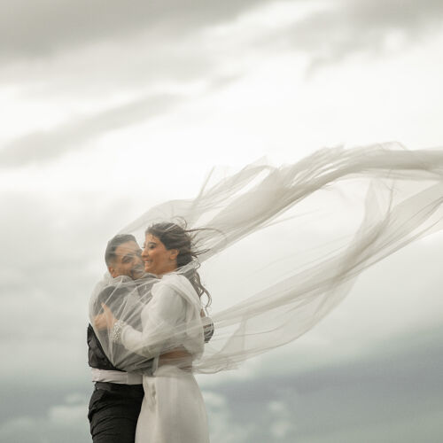 Hääkuva: groom holding bride, flowing veil in wind, couple embracing outdoors, overcast sky backdrop, romantic windswept moment, misty mountain setting | Giorgi Mikeladze, Tbilisi, Georgia