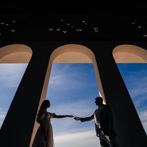 Wedding photo: couple holding hands under arches, silhouetted figures against sky, blue sky framed by columns, bride and groom backlit, architectural frames overhead | Stefan Liviu, București, Romania