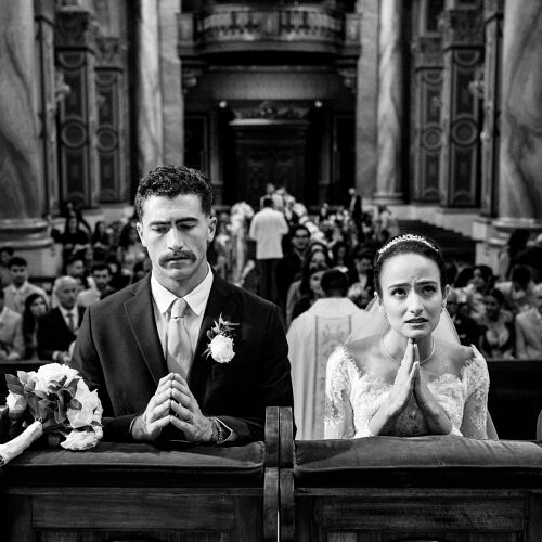 Bryllupsfoto: bride and groom kneeling, ornate church interior, black and white ceremony, formal wedding attire, crowded pews behind couple | Danilo &amp; Cris Almeida, Mogi das Cruzes, Brasilien