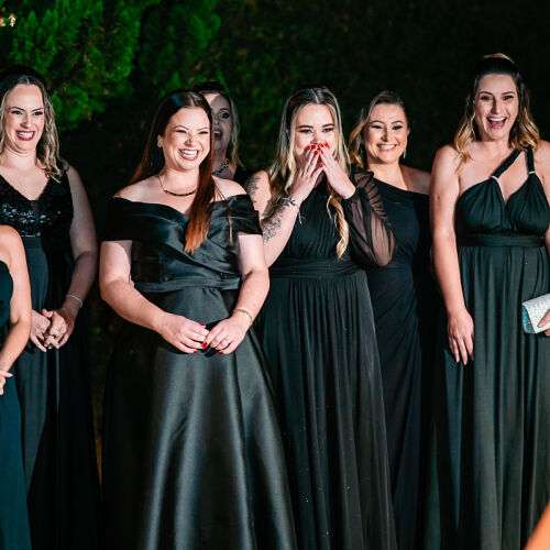 Wedding photo: bridesmaids in black dresses, nighttime outdoor event, women laughing together, green foliage backdrop, formal evening celebration | Diego Pereira Da Silveira, Tijucas, Brazil