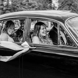 Bryllupsfoto: bride in vintage car, women laughing through window, classic automobile wedding, black and white portrait, natural daylight outdoor setting | Mischa Baettig, Steinhausen, Sveits