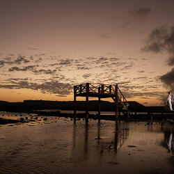 Wedding photo: couple walking on pier, sunset over water, wooden boardwalk at dusk, golden hour light, silhouetted figures by water, dramatic cloudy sky | Nestor G Zavarce, Willemstad, Curaçao