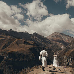 Wedding photo: bride and groom mountain valley, couple in white clothing mountains, alpine landscape wedding day, dramatic clouds mountain peaks | Giorgi Mikeladze, Tbilisi, Georgia