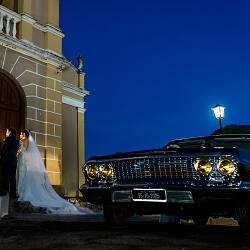 結婚式の写真: bride and groom at church door, vintage car with headlights on, twilight blue sky, couple in wedding attire, classic convertible at night | Flávio Roberto, Taubaté, ブラジル