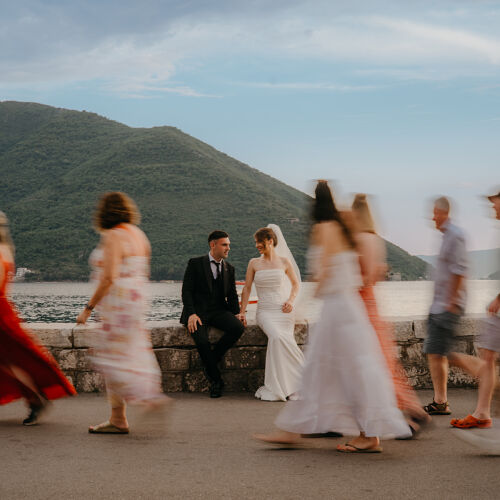 Wedding photo: bride and groom by water, guests running in motion blur, mountain backdrop, golden hour light, waterfront wedding celebration | Rale Petronijevic, Zlatibor, Serbia