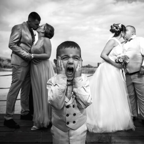 תמונת חתונה: child shouting at camera, couple kissing in background, waterside wedding party, black and white portrait, overcast sky, wooden dock setting | Plamen Petkov, Kazanlak, בולגריה