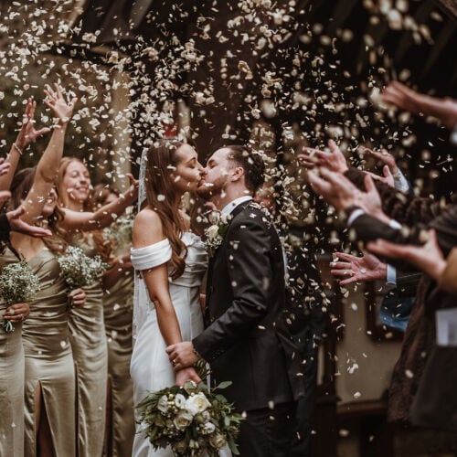 Düğün fotoğrafı: bride and groom kissing, confetti falling, wedding guests celebrating, off-shoulder white dress, black suit groom, indoor wooden venue | James Hatter, London, Birleşik Krallık