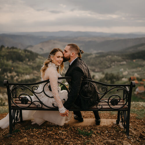 Wedding photo: bride and groom on bench, couple holding hands overlooking valley, ornate bench hillside, rolling hills landscape, overcast sky countryside | Rale Petronijevic, Zlatibor, Serbia