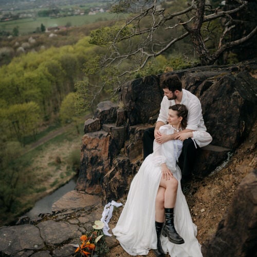 Zdjęcie ślubne: couple on rocky cliff, bride in white dress, groom embracing bride, valley landscape below, wildflowers on rocks, overcast daylight | Marie Veselá, Praha, Czechy