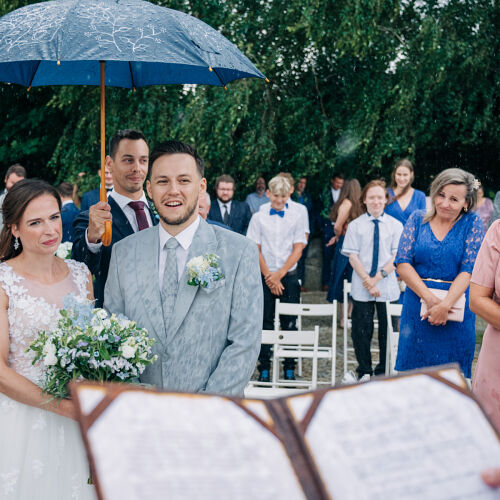 Zdjęcie ślubne: bride and groom smiling, outdoor garden wedding ceremony, blue umbrella in rain, guests seated watching, groom in gray suit, bride in white lace dress | Petr Hrubeš, Brno, Czechy