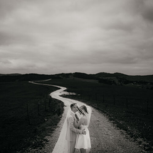 Wedding photo: couple kissing on rural road, winding river landscape, overcast sky, bride in white veil, groom embracing bride, moody countryside setting | Rale Petronijevic, Zlatibor, Serbia