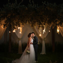 結婚式の写真: bride and groom embracing, nighttime garden wedding, hanging greenery and lanterns, warm golden lighting, couple under floral canopy | Vitor Oliveira, Teixeira de Freitas, ブラジル