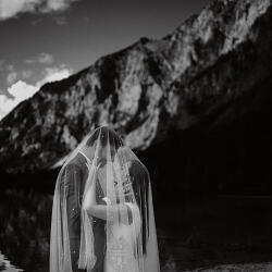 Wedding photo: bride in veil mountains, black and white wedding, mountain lake backdrop, dramatic mountain landscape, bride facing mountains, forest and peaks behind | Peter Schulz, Graz, Austria