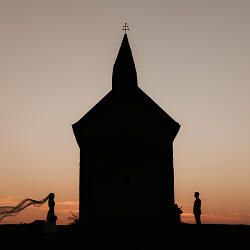 Zdjęcie ślubne: silhouette couple at sunset, bride with flowing veil, church building backlit, golden hour glow, two figures standing together | Robert Gálik, Bratislava, Słowacja