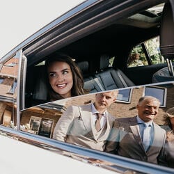 Wedding photo: woman smiling in car window, wedding guests in vehicle, brick building backdrop, daytime outdoor setting, group in formal attire | Helen Navajas, Amsterdam, Netherlands