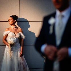 Svadobná fotografia: bride in white gown, off-shoulder wedding dress, dramatic shadow on wall, groom blurred in background, golden hour lighting | Andrei Hurduc, Vaslui, Rumunsko