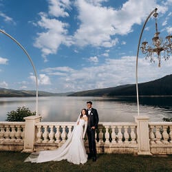 Esküvői fotó: bride and groom by lake, ornate chandelier arch, formal wedding attire, lakeside balustrade setting, forested mountains backdrop | Constantin Plugari, Chisinau, Moldova
