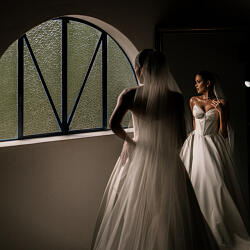 結婚式の写真: bride in white gown, dramatic window light, bride viewing mirror reflection, flowing pleated dress, dark interior setting, contemplative bridal moment | Eric Corbacho, São Paulo, ブラジル